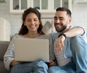 Image of a young, joyful couple sitting on a sofa, gazing at a laptop computer together